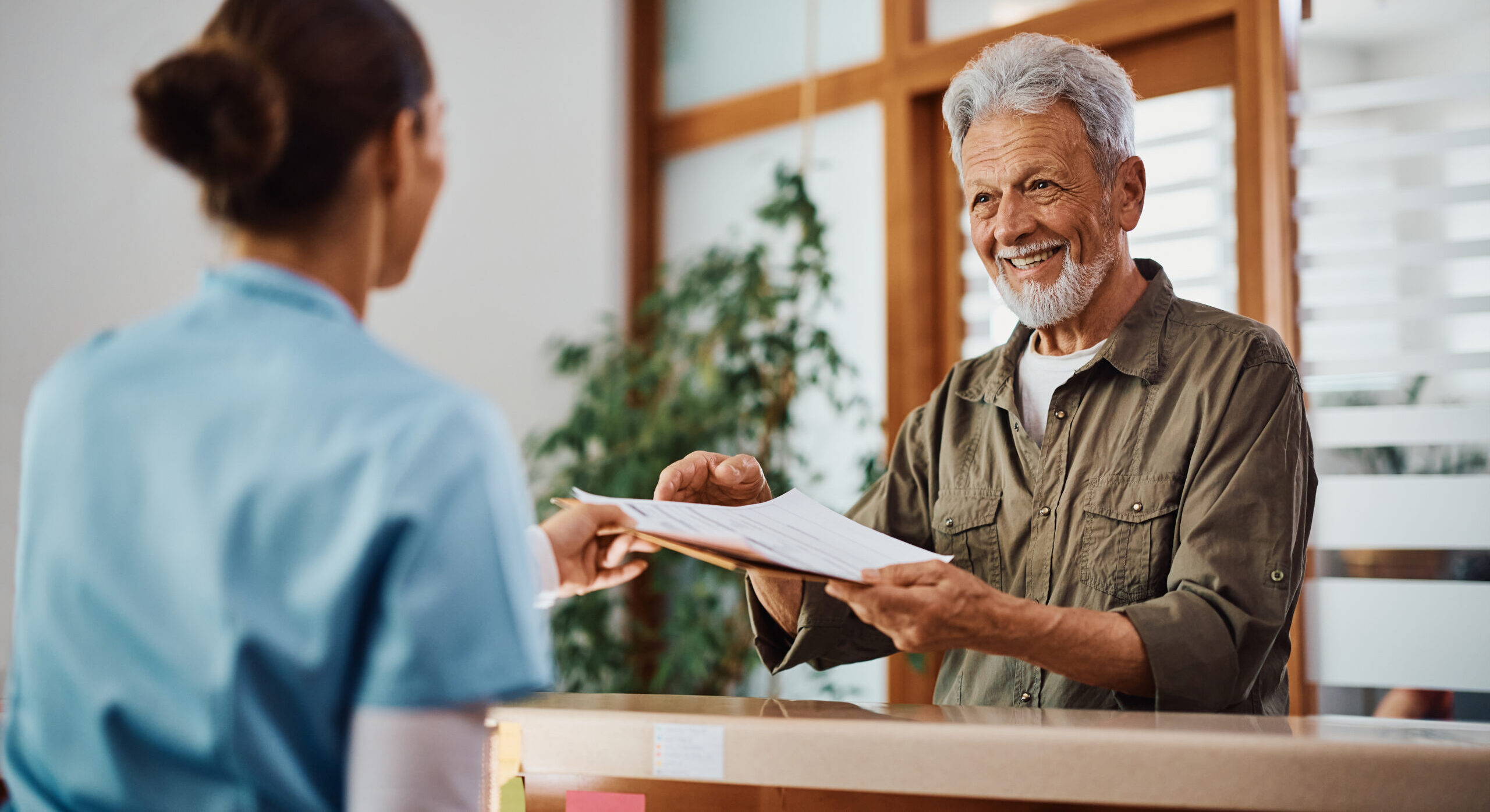 Happy senior man receiving his medical documents from nurse at reception desk at doctor's office.